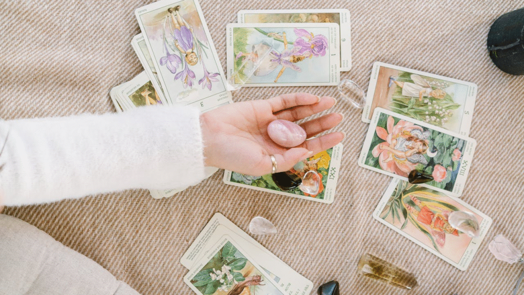 A woman’s hand holding a rose quartz crystal above a spread of illustrated tarot cards on a beige woven surface, symbolizing intuition, mindfulness, and modern spirituality.