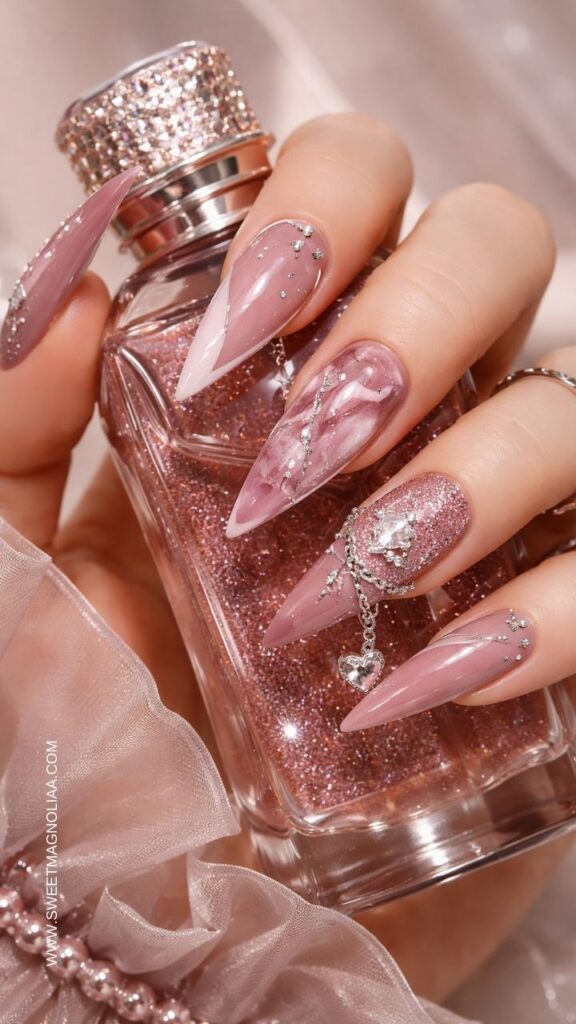 Blush pink stiletto nails with rhinestones, glitter accents, marble design, and a silver chain heart charm detail creating a glamorous luxury manicure look.