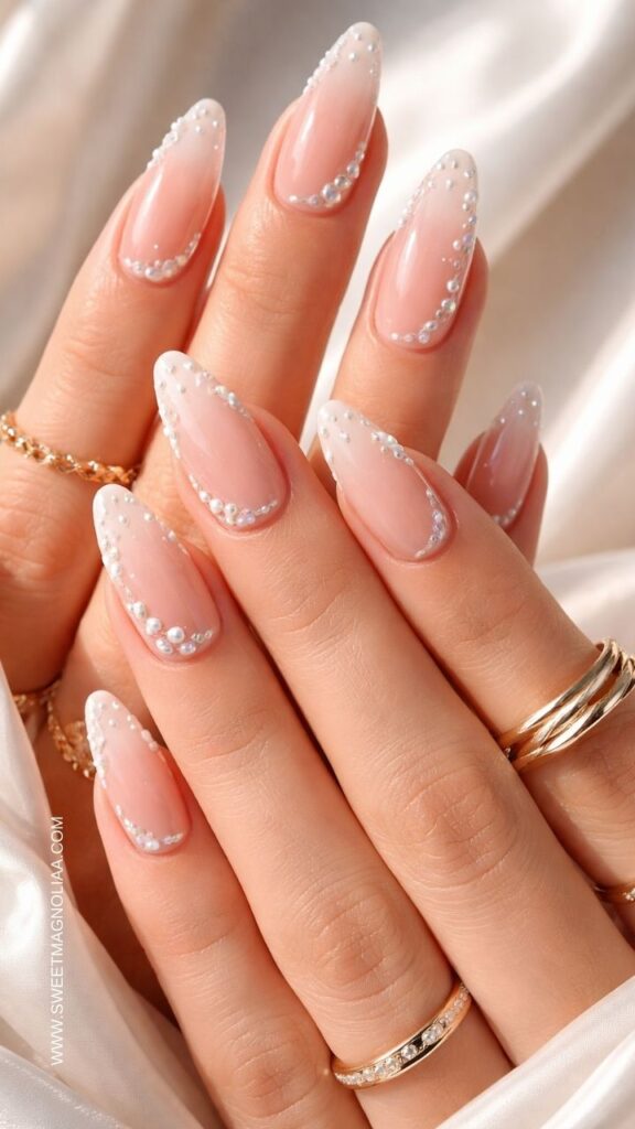 Soft pink almond nails with sheer French tips decorated with tiny pearl accents, elegant glossy manicure styled with gold rings, perfect bridal pink nail design inspiration.