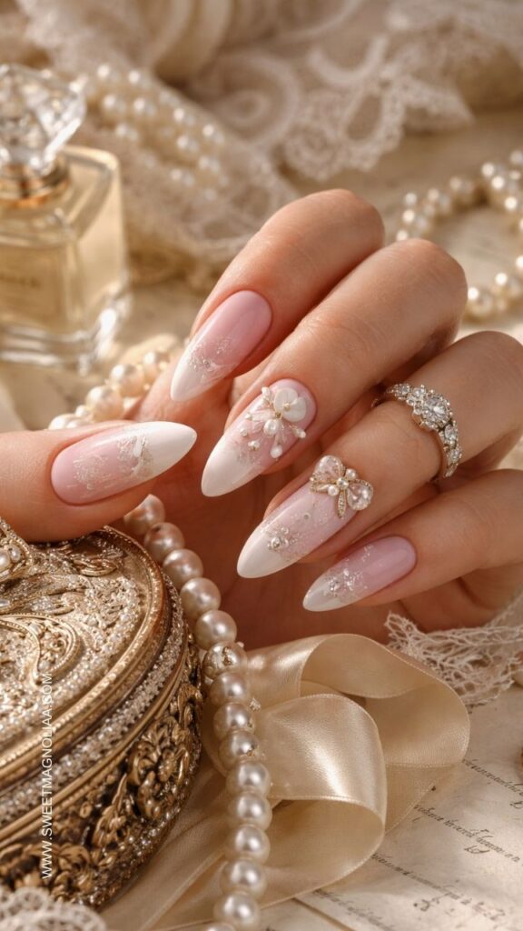 Blush pink almond nails with pearl flower accents, crystal bow charm, lace details, and soft shimmer creating an elegant romantic bridal manicure.