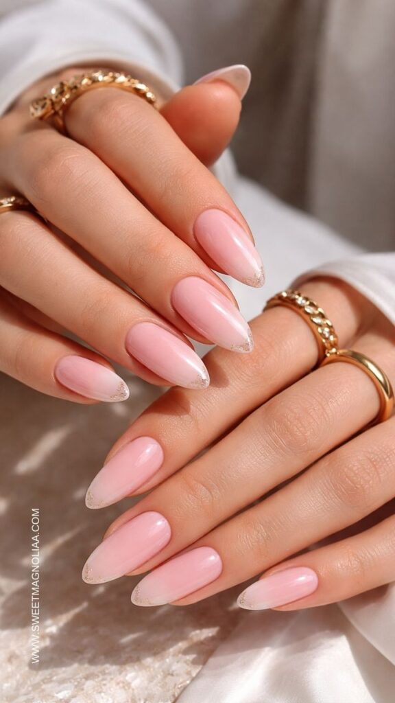 Soft pink almond nails with glossy finish and delicate glitter French tips, elegant minimalist manicure styled with gold rings, perfect for classy pink nail designs.