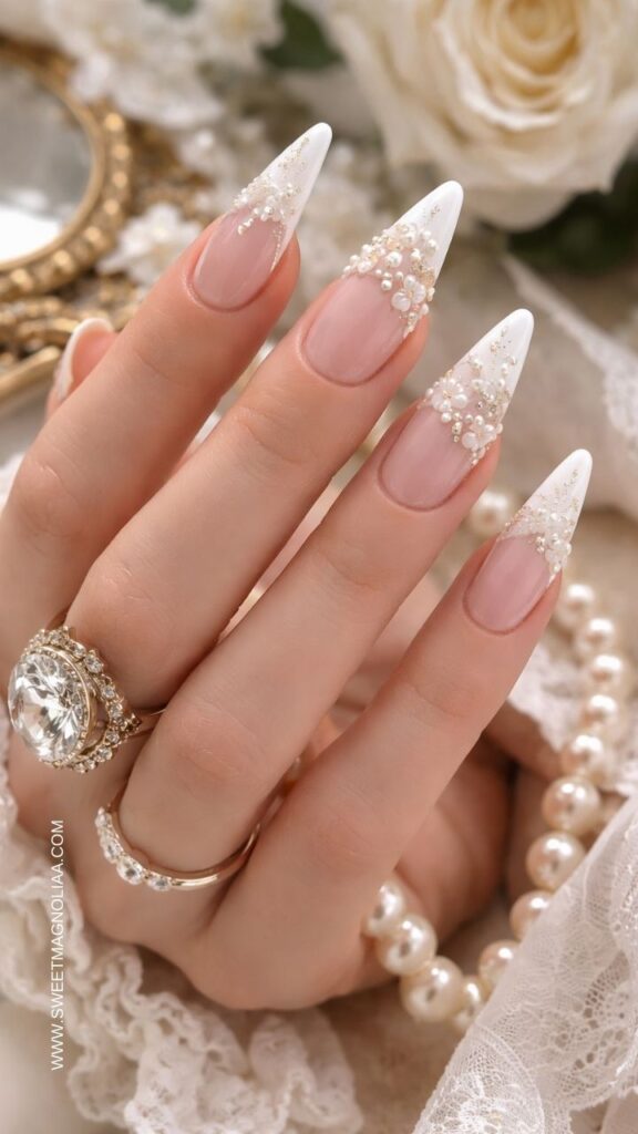 Blush pink almond nails with white French tips, pearl clusters, and crystal accents creating an elegant bridal inspired manicure with delicate luxury detail.