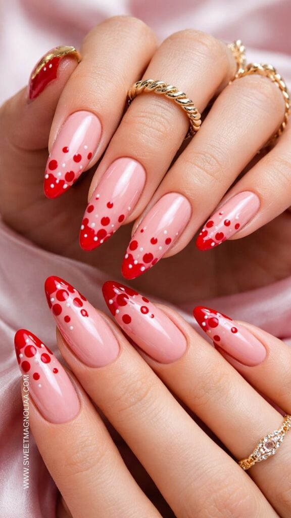 Glossy pink almond nails with red French tips and playful polka dot accents on a sheer blush base, stylish manicure with gold rings, perfect trendy pink nail design idea.