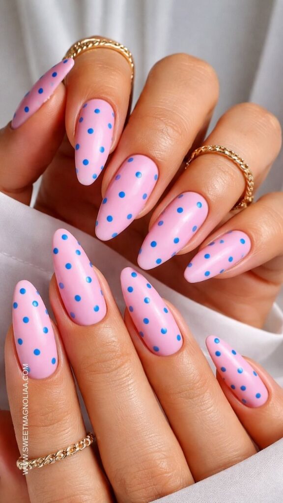 Glossy soft pink almond nails with small blue polka dots on each nail, playful pastel manicure styled with gold rings, cute retro inspired nail design idea