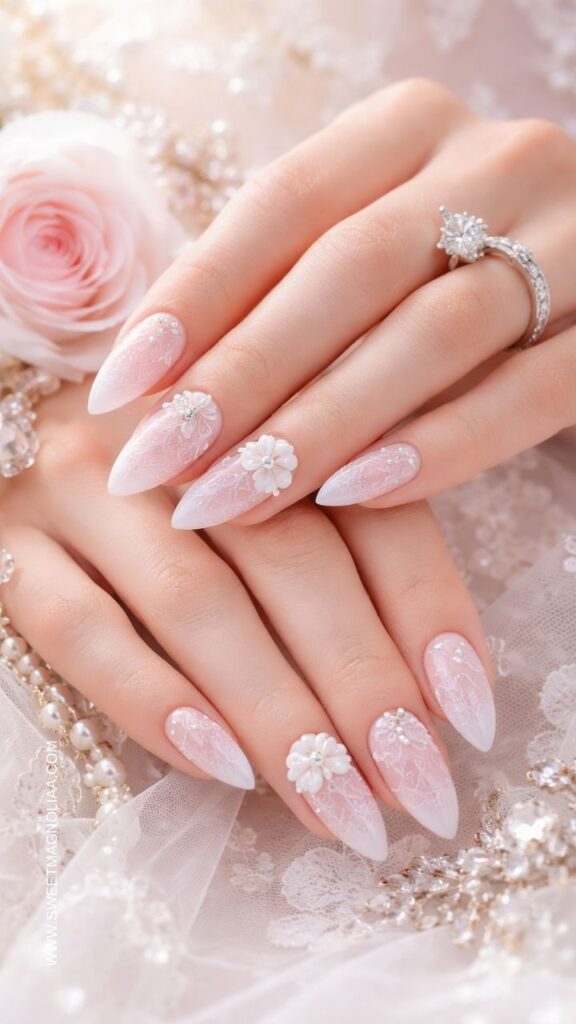 Blush pink ombre almond nails with delicate white lace patterns and small 3D floral accents creating an elegant romantic bridal manicure.