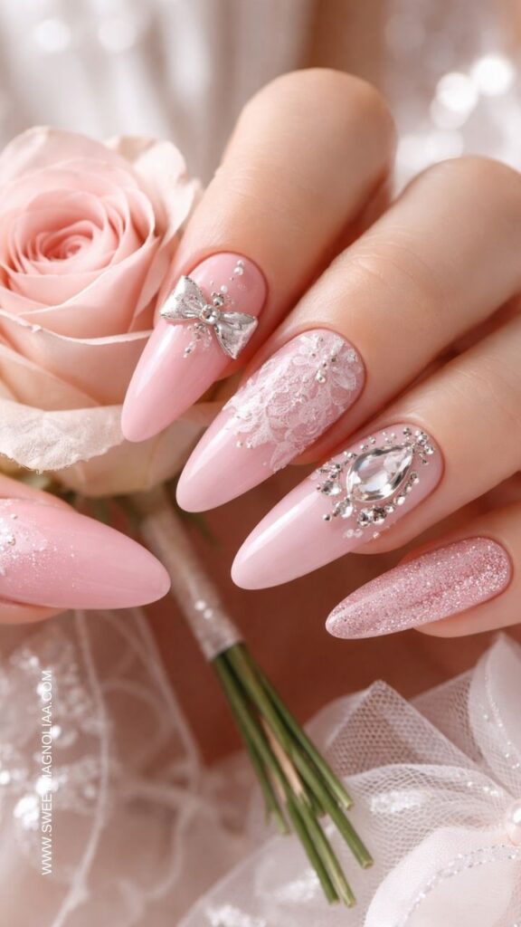 Soft pink almond nails with rhinestones, lace nail art, glitter accent nail and silver bow charm, elegant pink nail designs styled with a rose and bridal details.