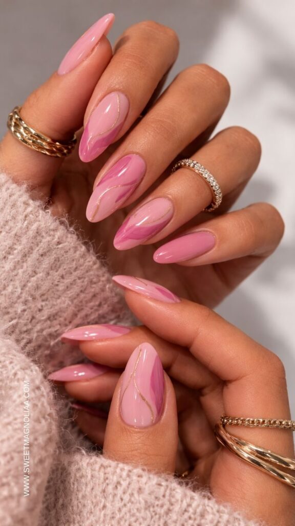 Glossy almond pink nail designs with marble swirl accents and thin gold lines, elegant feminine manicure styled with gold rings and soft pink sweater.