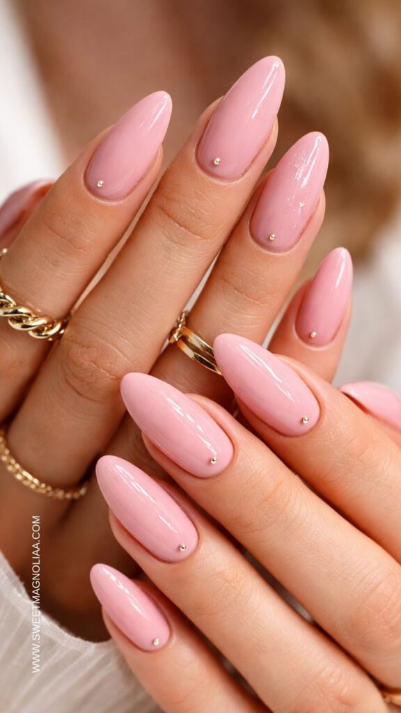 Glossy soft pink almond nails with delicate metallic stud accents on each nail, elegant minimalist manicure styled with gold rings, perfect classy pink nail design idea.